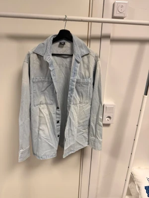 Ljusblå jeansjacka med Los Angeles-tryck - Säljer en ljusblå jeansjacka från VRS med stora bröstfickor och knappar framtill. På ryggen står det 'Los Angeles' i vit text. Jackan har klassisk krage och lång ärm, perfekt för dig som gillar streetstyle.