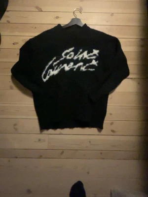 Svart stickad Saint Laurent tröja - Säljer en svart stickad tröja från Saint Laurent med vit, grafisk text på bröstet. Tröjan har lång ärm och en loose passform som ger en avslappnad vibe. Perfekt för dig som vill ha något trendigt och bekvämt. Lappar saknas då det är bortklippta och använt länge.  Passar som xs
