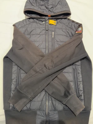 Parajumpers fleece & pufferjacka S - Säljer en grå Parajumpers hoodie-jacka i storlek S. Jackan har quiltad pufferfront, mjuka fleecesidor och ärmar, huva, dragkedja och ikonisk gul detalj vid halsen. Märkets patch på ärmen och flera praktiska fickor med dragkedja.