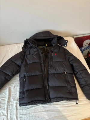 Svart pufferjacka från Polo Ralph Lauren - Svart pufferjacka från Polo Ralph Lauren med huva och två sidofickor med dragkedja. Jackan har ett rött Polo-logo broderat på bröstet och quiltad design. Perfekt för kalla dagar och har en snygg, modern look.