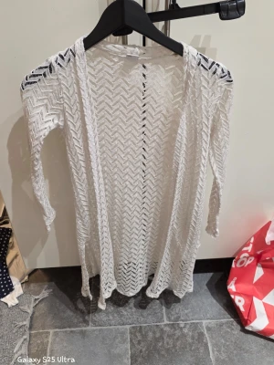 Vit lång kofta från Lindex - Säljer en vit, lång och öppen kofta från Lindex i storlek 134/140. Koftan är tillverkad i 100% polyester och har ett snyggt virkat mönster med långa ärmar. Perfekt att slänga över en topp för en chill look.
