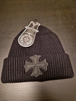 Svart mössa Chrome Hearts - Säljer en svart ribbad mössa från Chrome Hearts med ett stort kors i läder framtill och en metallknapp med logga på sidan. Mössan är i tjockt, stickat material och har en bred uppvikt kant. Perfekt accessoar för dig som gillar streetwear och exklusiva detaljer.