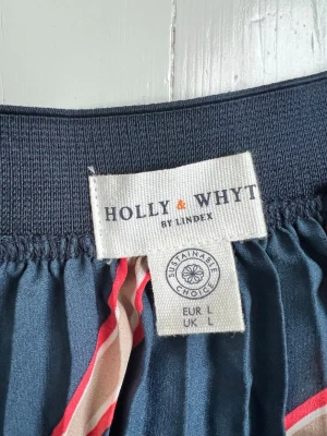 Randig plisserad kjol från Holly & Whyte - Säljer en marinblå plisserad långkjol från Holly & Whyte by Lindex i storlek L. Kjolen har diagonala ränder i rött, vitt och beige och ett elastiskt midjeband. Materialet känns lätt och flowy, perfekt för dig som gillar en färgstark och cool look.