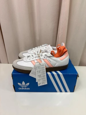 Adidas Samba vit och ljusrosa - Adidas Samba sneakers i vitt skinn med ljusrosa detaljer och klassiska tre ränder på sidan. Brun platt gummisula och rund tå. Skon har snörning och kommer med extra orange skosnören. Perfekt för dig som gillar retrostil och snygga färgkombinationer.
