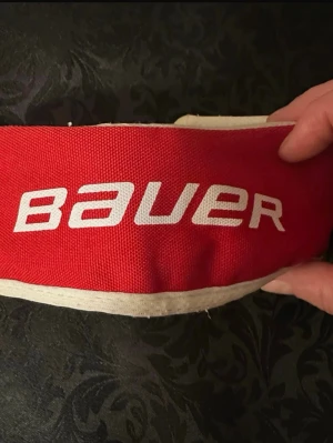 Röd Bauer skridskoskydd 26-35cm - Säljer ett rött skridskoskydd från Bauer med vit kant och tydlig logga. Passar skridskor med längd mellan 26 och 35 cm. Skyddet är gjort i slitstark textil och är perfekt för att hålla skenorna skyddade när du inte är på isen.