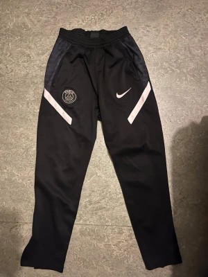 Svarta PSG x Nike träningsbyxor - Säljer ett par svarta träningsbyxor från Nike i samarbete med Paris Saint-Germain. Byxorna har vita detaljer på framsidan, PSG-logga och Nike Swoosh. De har elastisk midja och är gjorda i ett syntetmaterial som passar perfekt för sport och träning.