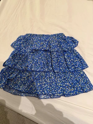 Blommig blå volangkjol från TUD101 - Säljer en blå kortkjol från TUD101 med blommigt mönster i vitt och grönt. Kjolen har flera lager med volanger och resår i midjan för en bekväm passform. Perfekt för dig som gillar färg och mönster!