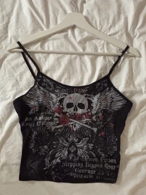 Svart linnetopp med dödskalletryck - Cool svart linnetopp med smala axelband och tryck av dödskalle, vingar och röda rosor framtill. Toppen har en edgy vibe med detaljerad grafik och text. Perfekt för dig som gillar alternativ stil och vill sticka ut.
