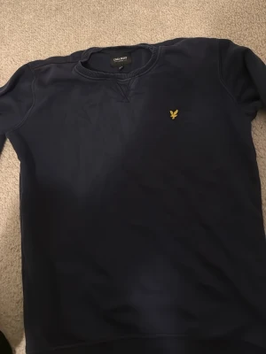 Marinblå sweatshirt från Lyle & Scott - Snygg marinblå sweatshirt från Lyle & Scott Junior med klassisk gul logga på bröstet. Tröjan har rund hals, ribbade muddar och lång ärm. Perfekt för dig som gillar stilrena och enkla plagg med en sportig touch.