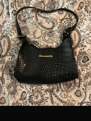 Svart väska  - Snygg svart axelväska i croco-mönstrat (blankt) material. Guldfärgad detalj framtill och dragkedja upptill. Perfekt storlek för vardag eller fest – får plats med mobil, nycklar och det viktigaste.