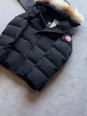 Svart pufferjacka från Canada Goose - Svart pufferjacka från Canada Goose med ikonisk röd och vit patch på ärmen. Jackan har huva med fluffig pälsdetalj, stora quiltade sektioner och knappar framtill. Perfekt för kalla dagar och har en riktigt snygg streetstil.