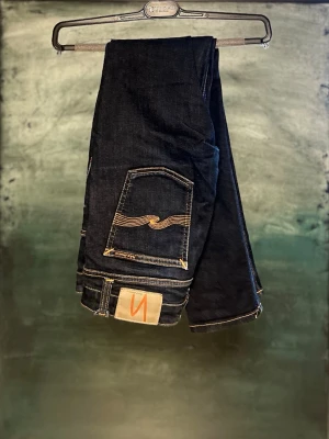 Nudie Jeans - Snygga mörkblå slim fit jeans från Nudie Jeans med klassisk femficksdesign och orangea sömmar. Jeansens bakfickor har Nudie Jeans ikoniska vågmönster och en patch med logga. Tillverkade i slitstark bomull med en slim fit passform och midwaist.