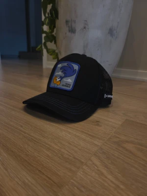 Svart Road Runner keps från Capslab - Säljer en svart keps från Capslab med Road Runner-tryck framtill. Kepsen har mesh på baksidan och broderad text på sidan. Framtill är det en blå och gul patch med Road Runner och moln. Perfekt för dig som gillar Looney Tunes✨️