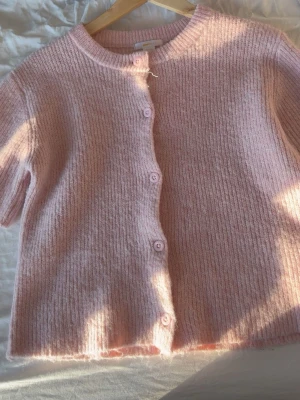 Ljusrosa stickad kofta från Gina Tricot - Superfin ljusrosa stickad kofta från Gina Tricot med rund hals och korta ärmar. Koftan har ribbad struktur och knäpps med matchande rosa knappar framtill. Perfekt att slänga över en topp för extra mysig stil.