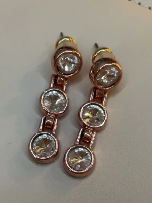 Guldiga örhängen med stenar - Säljer ett par snygga örhängen i guldig metall med tre runda, klara stenar på rad. Har bara använda ett fåtal gånger men har rengjort dom rejält och gör det igen inanna jag skickar iväg dom