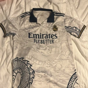 Real Madrid drakmönstrad fotbollströja - Säljer en vit Real Madrid fotbollströja från Adidas med detaljerat drakmönster i grått och mörkblått. Tröjan har korta ärmar, mörkblå krage och guldfärgade detaljer vid ärmslut. Perfekt för dig som vill sticka ut på planen eller bland kompisarna.