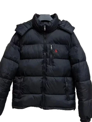 Svart pufferjacka från Polo Ralph Lauren - Svart pufferjacka från Polo Ralph Lauren med röd logga på bröstet. Jackan har hög krage, flera dragkedjeförsedda fickor och quiltad design. Materialet är polyester och jackan är perfekt för kalla dagar. Snygg och stilren look med långärmade ärmar och normal passform.