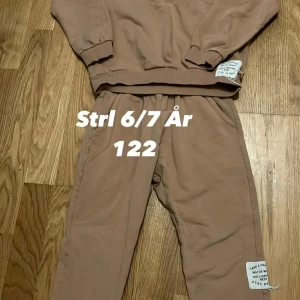 Beiga mjukisbyxor barn 122 - Mysiga beiga mjukisbyxor för barn i storlek 122. Byxorna har resår i midjan och muddar vid bensluten samt en vit patch med text på ena benet. Perfekta för chill dagar hemma eller till skolan.