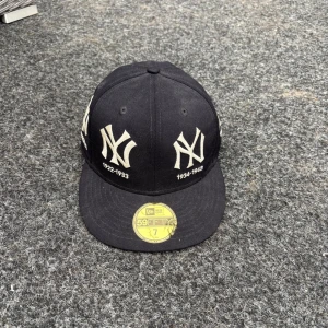 Svart New Era Yankees keps 59FIFTY - Snygg svart New Era 59FIFTY keps med New York Yankees loggor broderade runt hela kepsen och årtal under varje logga. Platt skärm och klassisk passform. Materialet är 100% ull och kepsen har en gul etikett på skärmen. Perfekt för dig som gillar streetwear och baseball.