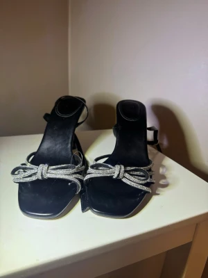 Svarta klack-sandaler med strassrosett - Säljer ett par svarta sandaler från H&M med smal hög klack och fyrkantig tå. Skorna har glittrande strassdetaljer i form av en rosett framtill och tunna remmar runt ankeln. Materialet är syntet med en elegant finish, perfekt för att lyfta din outfit.