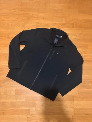 Svart vindjacka från Polo Ralph Lauren - Svart vindjacka från Polo Ralph Lauren med stilren design och diskret logga på bröstet. Jackan har hel dragkedja framtill, två sidofickor med dragkedja och en bröstficka med dragkedja. Materialet är syntet och jackan har långärmade ärmar.