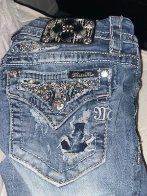 Miss Me blå jeans med strass och broderi - Snygga blå jeans från Miss Me med bootcut-snitt och coola detaljer som strass, broderier och slitningar. Fickorna har glittrande dekorationer och metallknappar med mönster. Perfekt för dig som gillar statement-jeans med unik look. Lowwaist bootcut i storlek W25, jättebra skick