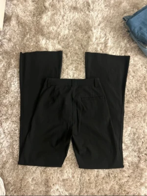 Svarta bootcut kostymbyxor - Snygga svarta kostymbyxor med bootcut-snitt och klassiska fickor både fram och bak. Byxorna har hällor för bälte och är tillverkade i ett mjukt och följsamt material som ger en clean och stilren look. Perfekta för dig som gillar en lite vidare passform nertill.