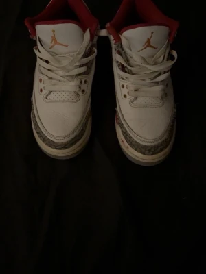 Nike Air Jordan 3 Retro Vit/Röd - Nike Air Jordan 3 Retro sneakers i vitt skinn med röda och grå detaljer. Klassisk siluett med snörning, rund tå och ikoniskt elefantmönster runt tå och häl. Röd insida och Jumpman-logga på plös och häl. Snygga detaljer och retrovibbar. Bra skick bara skitiga