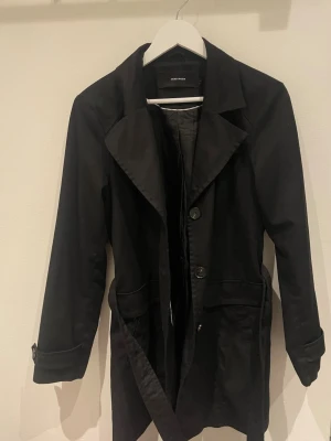 Svart trenchcoat från Vero Moda - Säljer en svart trenchcoat från Vero Moda i storlek L. Jackan har klassisk krage, knappar framtill och bälte i midjan för snygg passform. Långa ärmar med detalj vid ärmslut och två fickor framtill. Perfekt för dig som vill ha en stilren och enkel jacka.