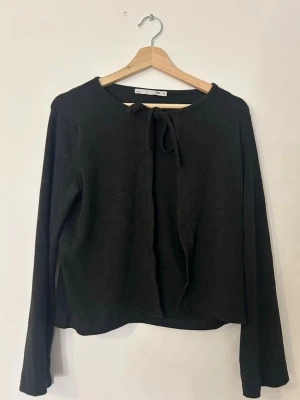 Svart kofta med knyt framtill - Mysig svart kofta från 1902 i mjukt material. Koftan har långa ärmar och en enkel knytning framtill vid halsen som ger en söt touch. Perfekt att slänga över en topp för en chill look.