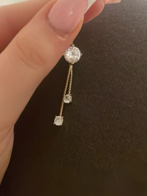 Guldig navelpiercing - Säljer en snygg navelpiercing i guld-look med tre glittrande stenar på tunna kedjor. Piercingen har en böjd stav och två hängande kedjor med små, klara stenar som ger extra bling. Perfekt för dig som vill ha något som sticker ut.