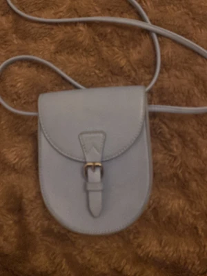 Ljusblå axelväska med spänne - Säljer en stilren ljusblå axelväska i skinnimitation med rundad form och ett snyggt spänne framtill. Man kan ha den crossbody eller bara vid axeln. den är knappt andvändts 