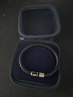 Svart flätat armband Tommy Hilfiger - Snyggt svart armband från Tommy Hilfiger med flätad design och ett stilrent metallspänne med logotypdetalj. Armbandet kommer i originaletui och har en modern look som passar perfekt till streetwear eller casual outfits. Längd cirka 21 cm.