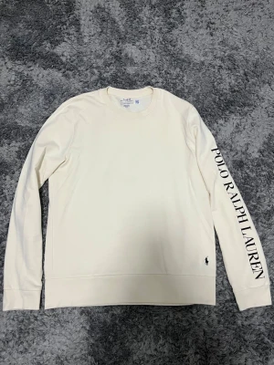Vit långärmad tröja Polo Ralph Lauren - Snygg vit långärmad tröja från Polo Ralph Lauren med svart logotyp broderad vid nederkanten och stor text på ena ärmen. Tröjan har rund hals och är gjord i mjuk bomull, perfekt för chill dagar. Passar dig som gillar stilrena och klassiska plagg.