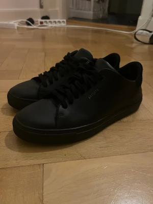 Svarta sneakers från Axel Arigato - Säljer ett par stilrena svarta sneakers från Axel Arigato i storlek 42. Skorna har en minimalistisk design med rund tå, platt sula och snörning. Tillverkade i skinn med diskret logga på sidan, sulan och hälen. Perfekta för dig som gillar cleana och moderna sneakers.