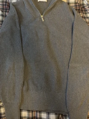 Grå ribbad stickad tröja med half zip - Säljer en grå stickad tröja med ribbad struktur och half zip framtill. Tröjan har långärmade och breda muddar vid ärmslut och nederkant. Perfekt för dig som gillar en enkel men snygg look med lite edge.