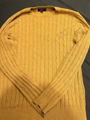 Gul kabelstickad tröja från Gant - Gul kabelstickad tröja från Gant med rund halsringning och diskret broderad logga på bröstet. Tröjan har långa ärmar och ribbade muddar vid ärmslut och nederkant. Perfekt för dig som vill ha en klassisk men färgstark look.