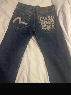 Evisu jeans med silverdetaljer - Unika blå jeans från Evisu med stora silvriga broderier på bakfickorna – ena med Evisu Genes Osaka-tryck och andra med klassiska måsen. Klassisk femficksmodell i jeansmaterial och raka ben. Perfekt för dig som vill sticka ut med streetwear-vibe. Jeansen är använda ca 5 gånger och har inga defekter förutom ytterst lite heelbite men inget man tänker på! Dessa jeans är väldigt sällsynta! Hör gärna av er angående fler bilder/ frågor! Skriv gärna innan köp☺️