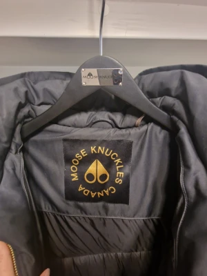 Svart lång dunjacka Moose Knuckles - Säljer en svart lång jacka från Moose Knuckles med guldfärgade detaljer och bälte i midjan. Dem stängs framtill med guldfärgad tvåvägsdragkedja. Bröstficka på vänster sida och två fickor framtill. Tyvärr råkade jag riva upp ca 4 cm längs ner på jackan som man kan sy ihop och det är inget som syns när man bär den. 