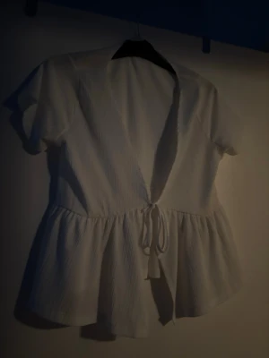 Vit omlottopp med knyt - Säljer en söt vit omlottopp från 157 i storlek XS. Toppen har korta ärmar, v-ringning och knyt framtill i midjan. Materialet är lätt och har en diskret struktur som ger en luftig känsla. Perfekt till jeans eller kjol för en fräsch look.