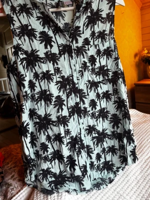 Ärmlös skjorta med palmmönster från Divided - Säljer en ärmlös skjorta från Divided med svart palmmönster på ljus bakgrund. Skjortan har knappar framtill och en bröstficka. Perfekt för varma dagar och ger en chill vibe till outfiten.