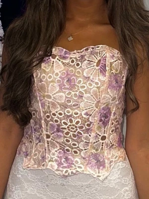 Blommig spetskorsett - Säljer en superfin korsettopp med blommigt spetsmönster i ljusa pastellfärger som lila och beige. Toppen är axelbandslös och har en snygg figurnära passform💕 har använts endast en gång!!