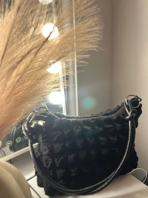 Svart axelväska med quiltad look - Säljer en svart axelväska med cool quiltad textur och bred axelrem i skinnimitation. Väskan har dragkedja upptill och snygga metall-detaljer vid remmens fästen. Perfekt storlek för det viktigaste och riktigt trendig form.