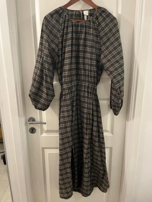 Rutig långklänning från H&M - Säljer en rutig långklänning från H&M i beige, grå och svart. Klänningen har lång ärm, rund halsringning och resår i midjan för en snygg siluett. Perfekt för dig som gillar en avslappnad men ändå stilsäker look.