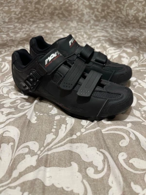 Svarta cykelskor från RDM - Säljer ett par svarta cykelskor från RDM med robust gummisula och SPD-klossar. Skorna har tre kardborreband och en spänne för extra stabilitet. Ovandelen är i syntetmaterial med meshpaneler för ventilation. Perfekt för dig som gillar att cykla och vill ha bra grepp.