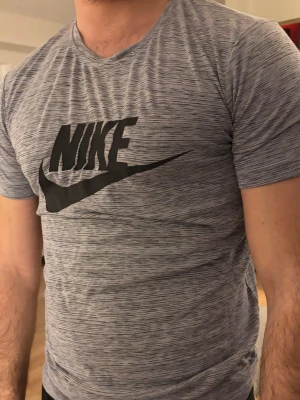 Grå Nike t-shirt med svart logga - Säljer en gråmelerad t-shirt från Nike med stor svart logga på bröstet. T-shirten har korta ärmar och är tillverkad i ett mjukt och lätt material som passar perfekt för träning eller chill. Slim fit för en snygg och sportig look.