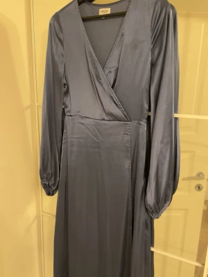 Marinblå omlottklänning från Bubbleroom - Snygg marinblå långklänning från Bubbleroom i glansigt material med omlott-design och v-ringad hals. Klänningen har långa, puffiga ärmar med resår vid handlederna och en elegant siluett som faller fint längs kroppen.