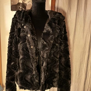 Svart pälsjacka från Vero Moda - Mysig svart pälsjacka från Vero Moda med huva och knappar framtill. Jackan har ett vågigt mönster i pälsen och är fodrad på insidan för extra komfort. Perfekt för dig som vill ha en varm och snygg jacka till kyliga dagar.