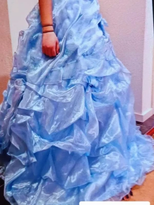 Ljusblå långklänning med volanger - Säljer en drömmig ljusblå långklänning med glansigt tyg och stora volanger. Klänningen har en riktigt prinsessig känsla och är perfekt för bal eller fest. Materialet är skimrande och ger ett lyxigt intryck. Passar dig som vill sticka ut och känna dig extra speciell.