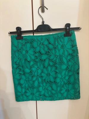 Grön blommig spetskjol - Snygg grön kortkjol med blommigt spetsmönster. Kjolen har en rak passform och är täckt av detaljerade blommor i spetsmaterial som ger en cool och unik look. Perfekt att matcha med en enkel topp för en trendig outfit.